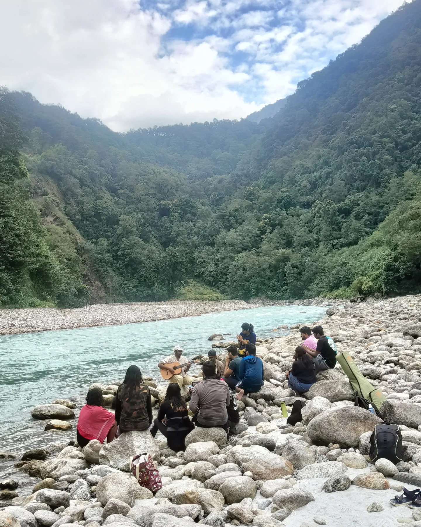 A group gathered on river rocks, one playing guitar, beside a Himalayan river in Sikkim.